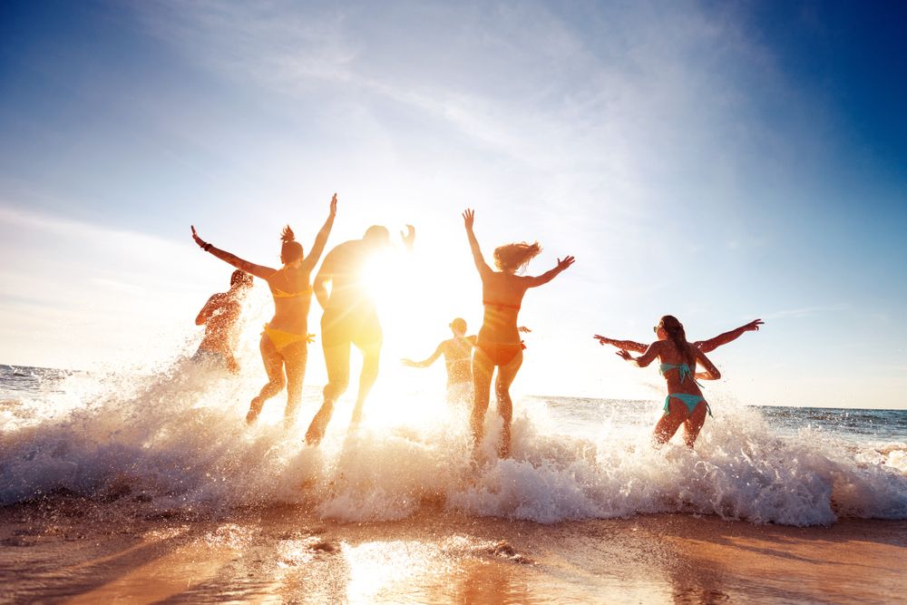 People jumping in the ocean with arms raised, sunlit beach, waves crashing, and bright sky.