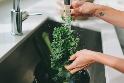 Hands rinsing kale leaves under a faucet in a stainless steel sink.