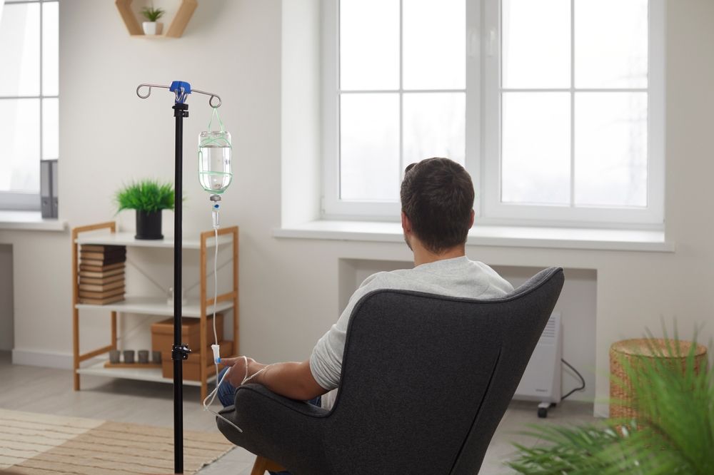 Person receiving IV treatment, seated in a chair, with a window and equipment in a home setting.