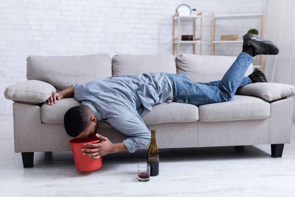Person passed out on a couch, head in a bucket, near a bottle of wine and a glass.