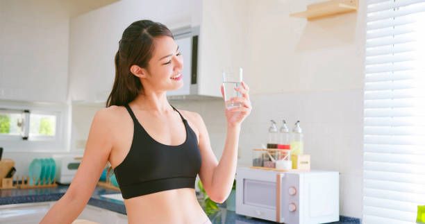 Woman in workout clothes holding glass of water in a kitchen, smiling.