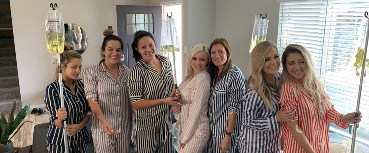 Seven women in pajamas pose together indoors, some holding IV poles.