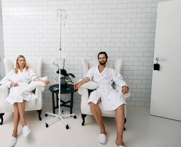Two people in white robes sit in chairs, receiving IV drips in a spa-like room.