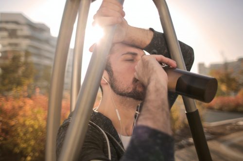 Man taking a drink from a bottle, outdoors. Sun shines through exercise equipment.