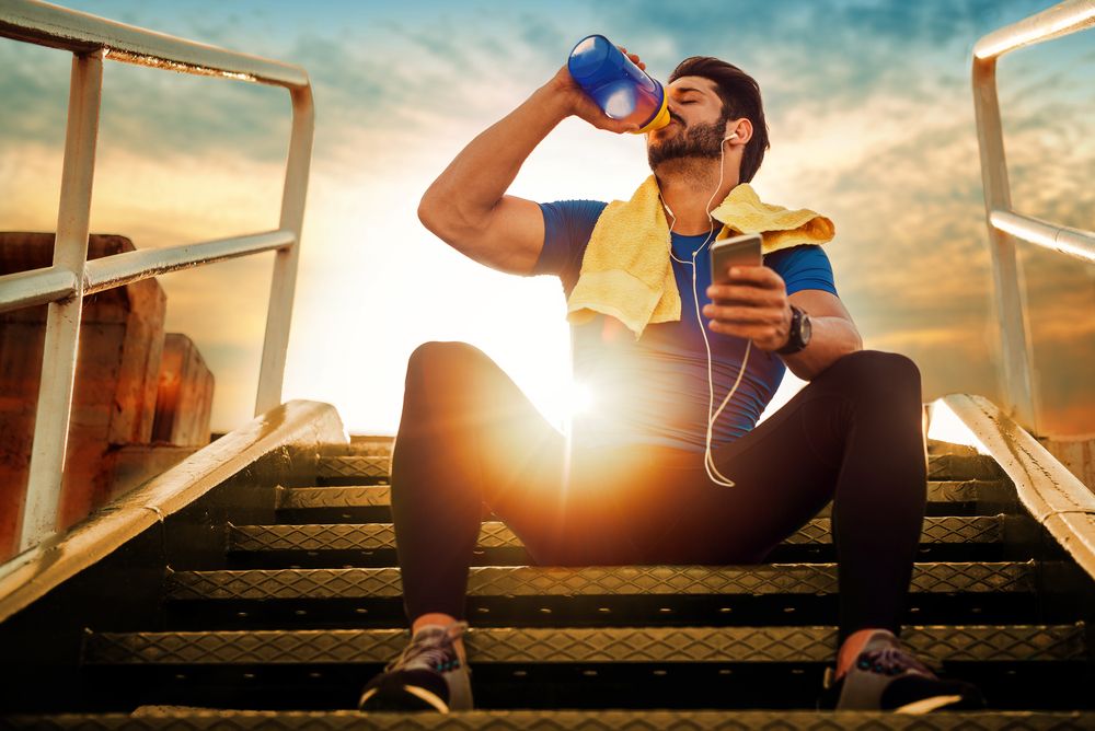 Man drinking from a water bottle while sitting on steps, with a towel around his neck; sunset.