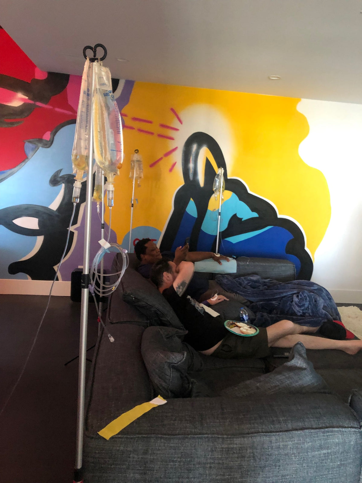 Person on couch receiving IV fluids, colorful mural backdrop.