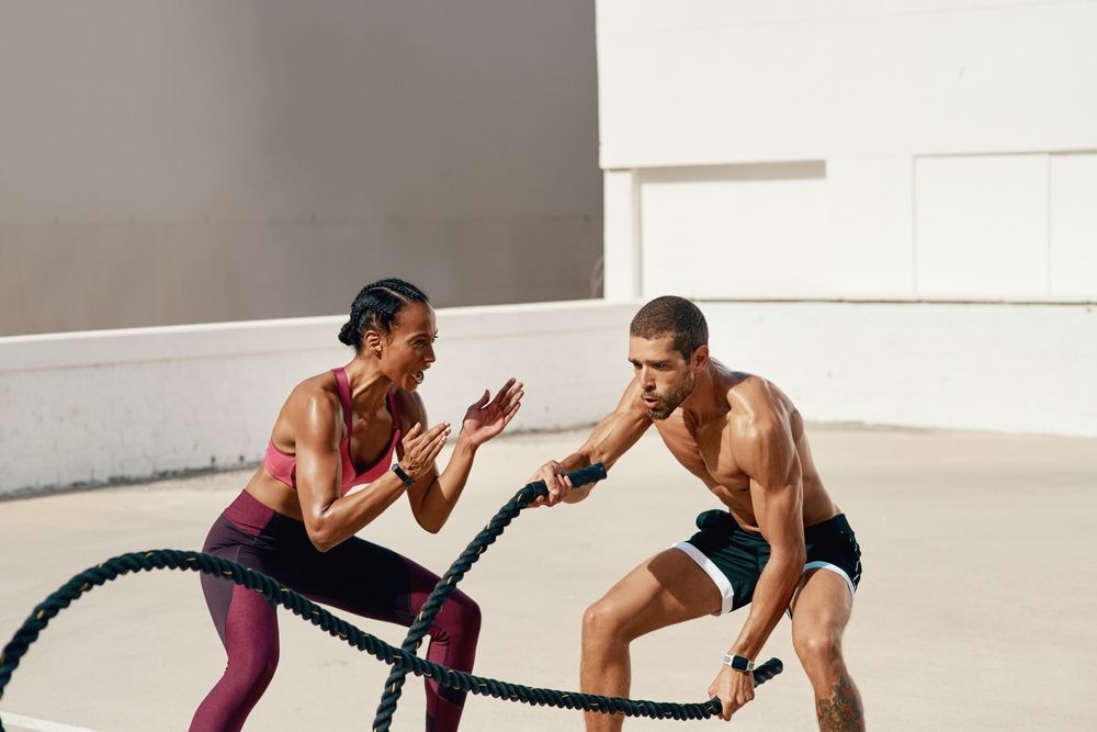 Man and woman doing battle rope exercises outdoors. Both are sweating.