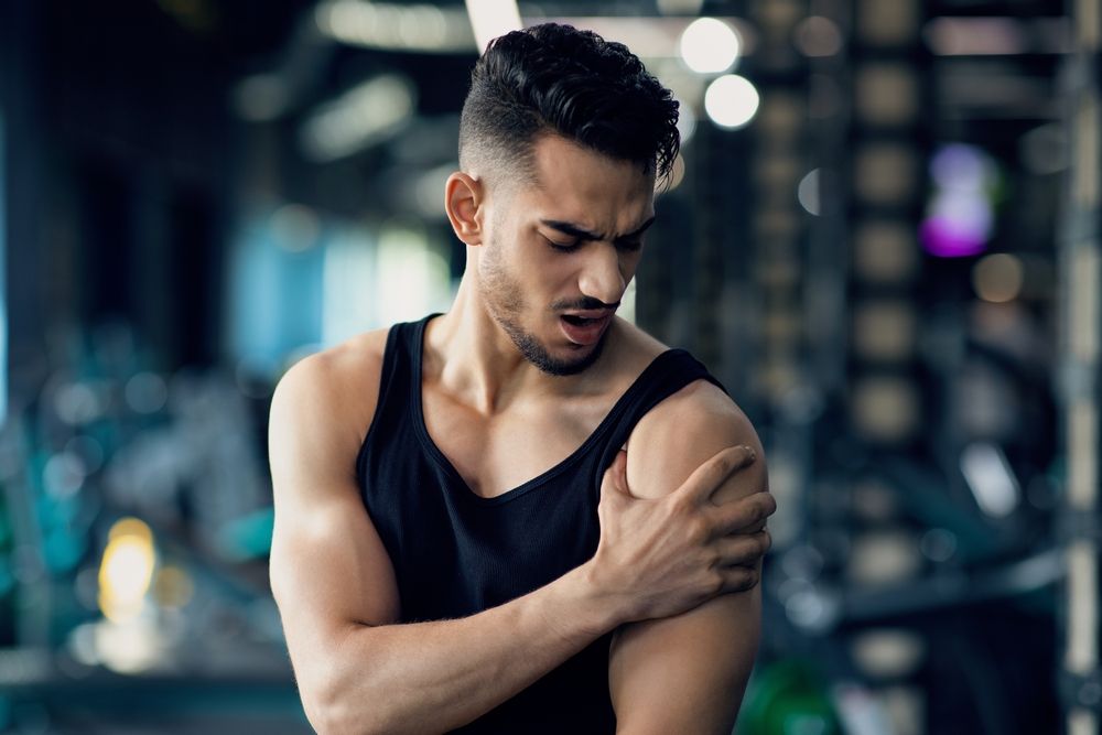 Man at gym clutching shoulder, grimacing in pain. Black tank top, dark hair, blurry background.