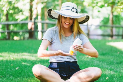 Woman wearing hat applies sunscreen to hand while sitting on grass.