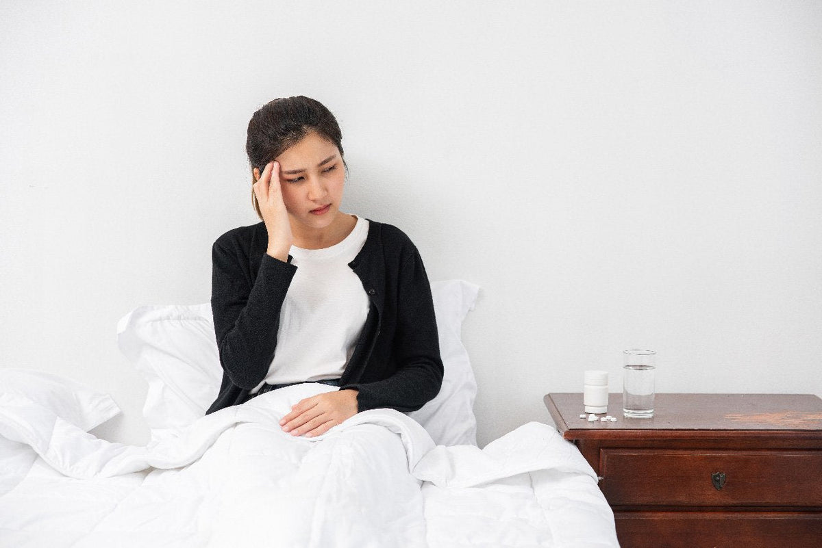 Woman in bed, holding head, appears to have a headache. Medication on nightstand.