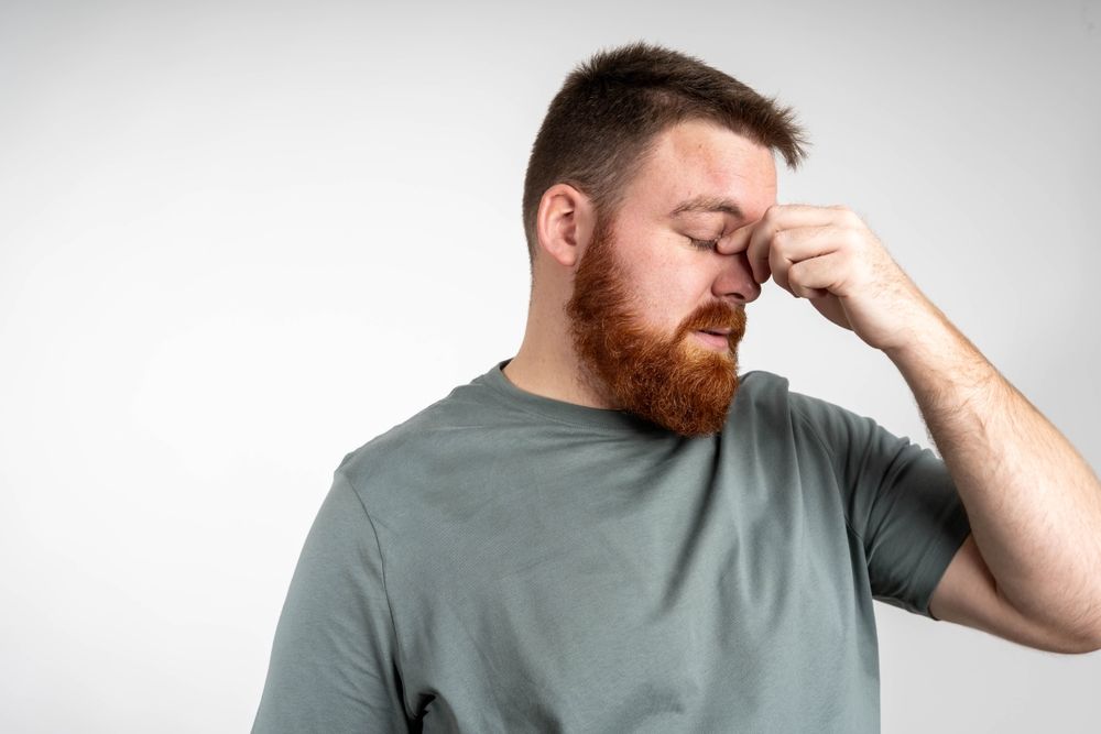 Man with red beard, wearing a grey shirt, pressing his fingers against the bridge of his nose, looking down with a pained expression.