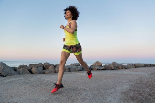 Woman running on a concrete path near the water, wearing a yellow top and patterned shorts.