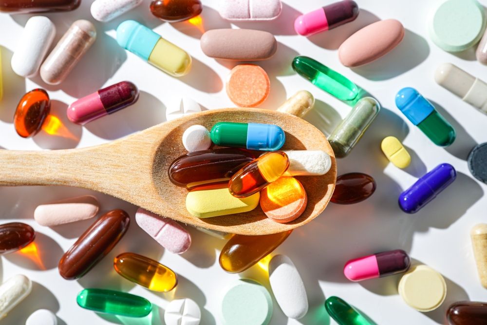Pills of various colors and shapes scattered on a white surface, with some in a wooden spoon.