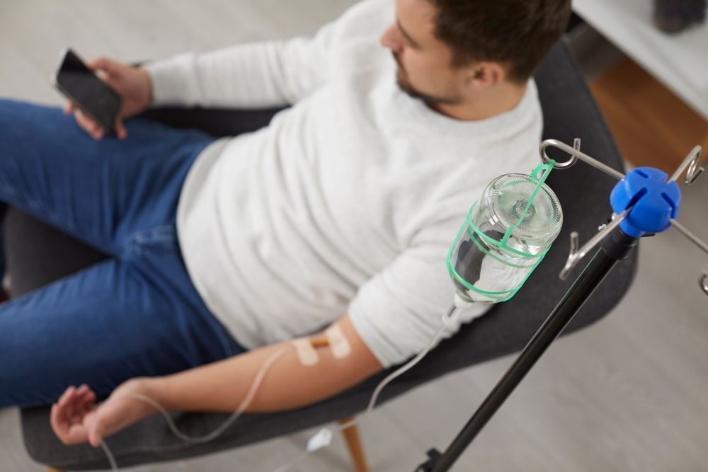 Man receiving IV fluids while sitting in a chair, holding phone.