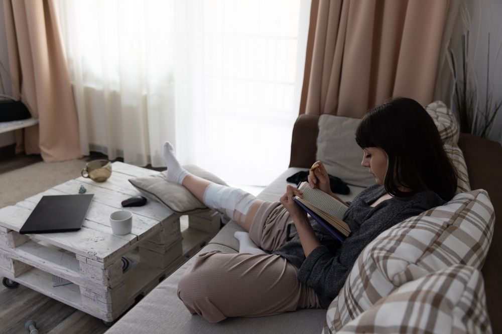Woman with leg in cast reads book on couch, laptop and coffee table nearby.