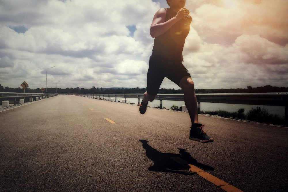 Runner in dark shorts and top leaps forward on a sunny road, casting a long shadow.