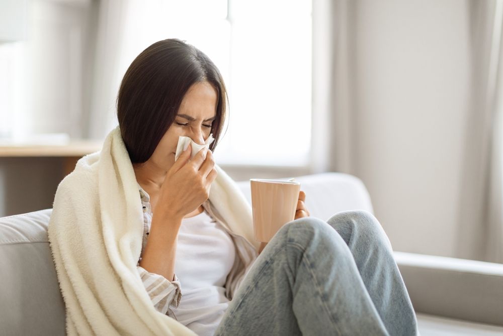 Woman on a couch blowing her nose with a tissue; holding a mug and wrapped in a blanket.
