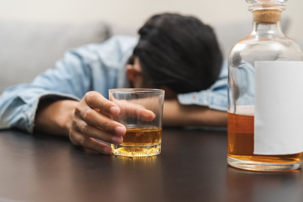Person slumped on table, reaching for glass of amber liquid. Bottle of alcohol nearby.