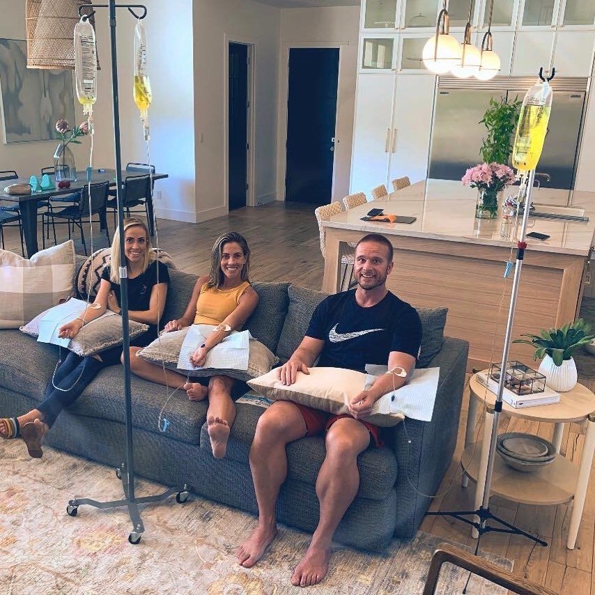 Three people receiving IV infusions on a couch in a living room. Bags hang from IV stands.