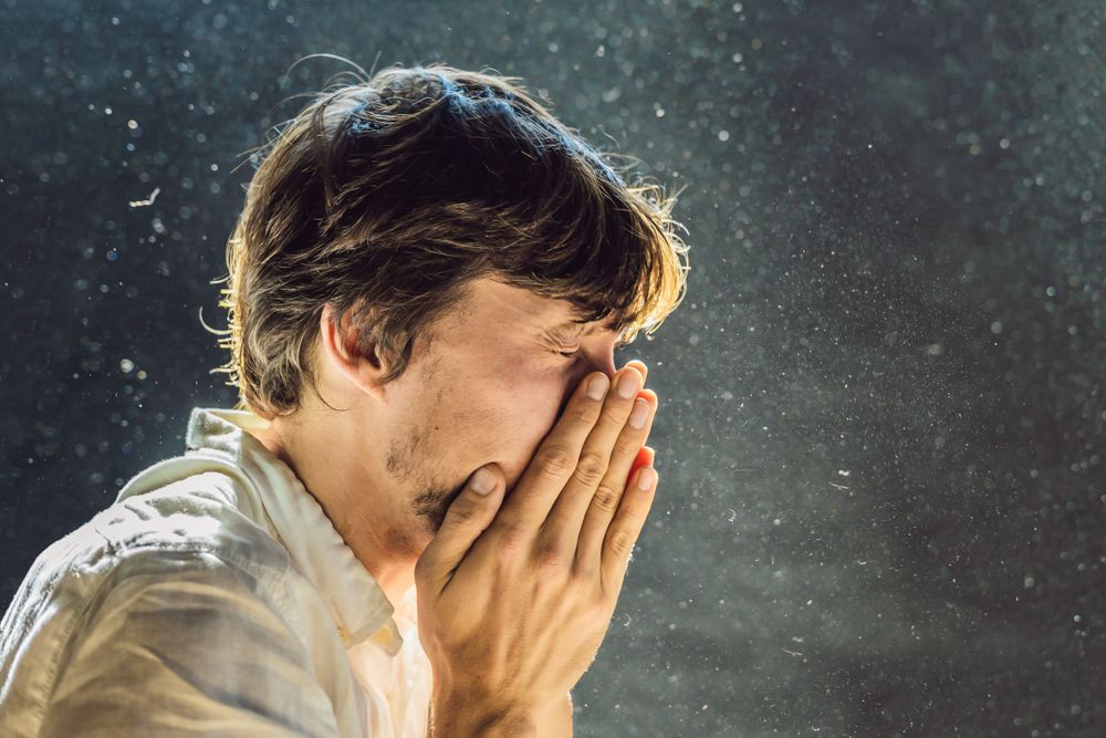 Man covering face, sneezing, with dust particles in air.