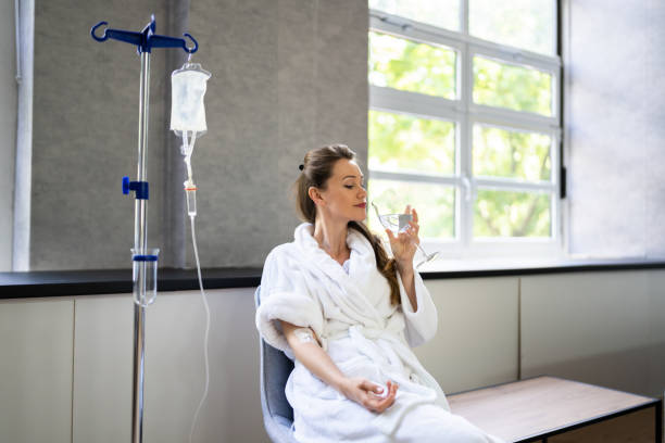 Woman in bathrobe receiving IV drip, drinking water by a window.