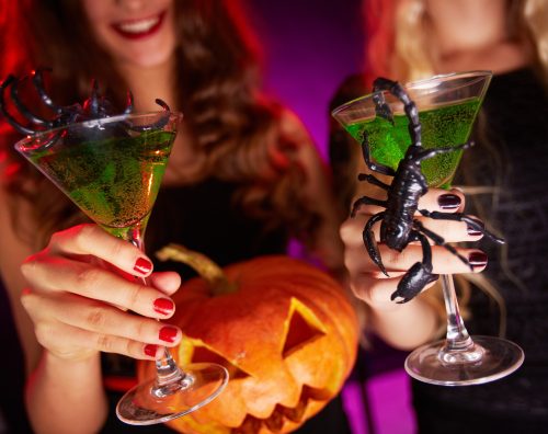 Two women holding green cocktails with plastic spiders, jack-o'-lantern, Halloween party.