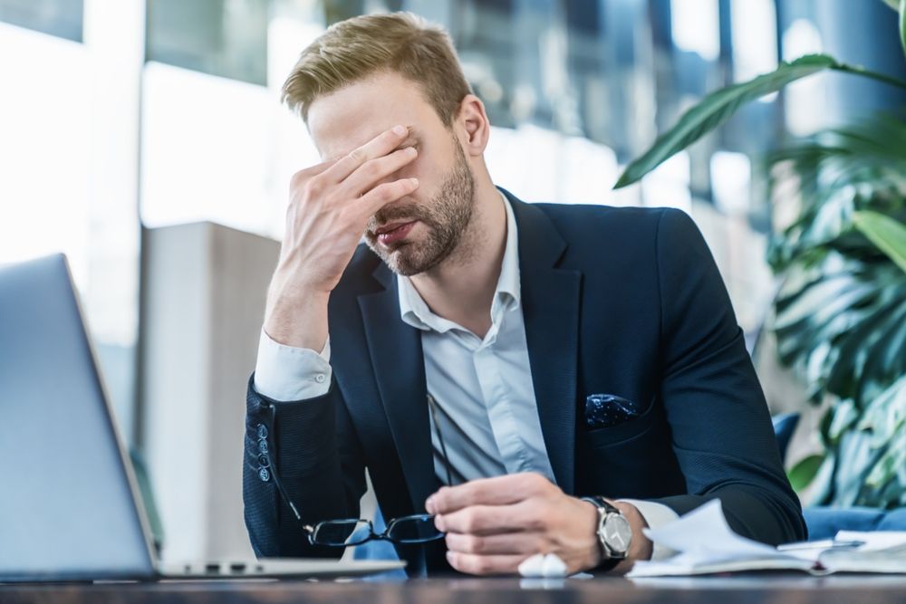 Man in suit, hand over face, appears stressed, laptop and paperwork in front.