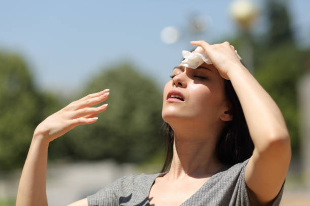 Woman wipes sweat from forehead in the sun, arms raised, looks distressed.