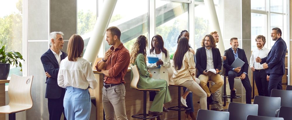 Group of people in an office space, some standing and talking, others seated on stools.