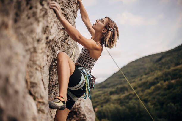 Woman rock climbing, reaching up with a hand on a rock face. She's wearing a harness and climbing shoes.