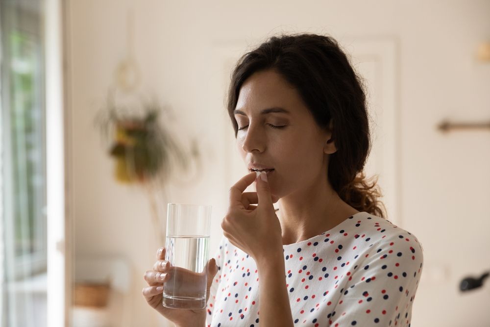 Woman taking a pill with a glass of water indoors, eyes closed.