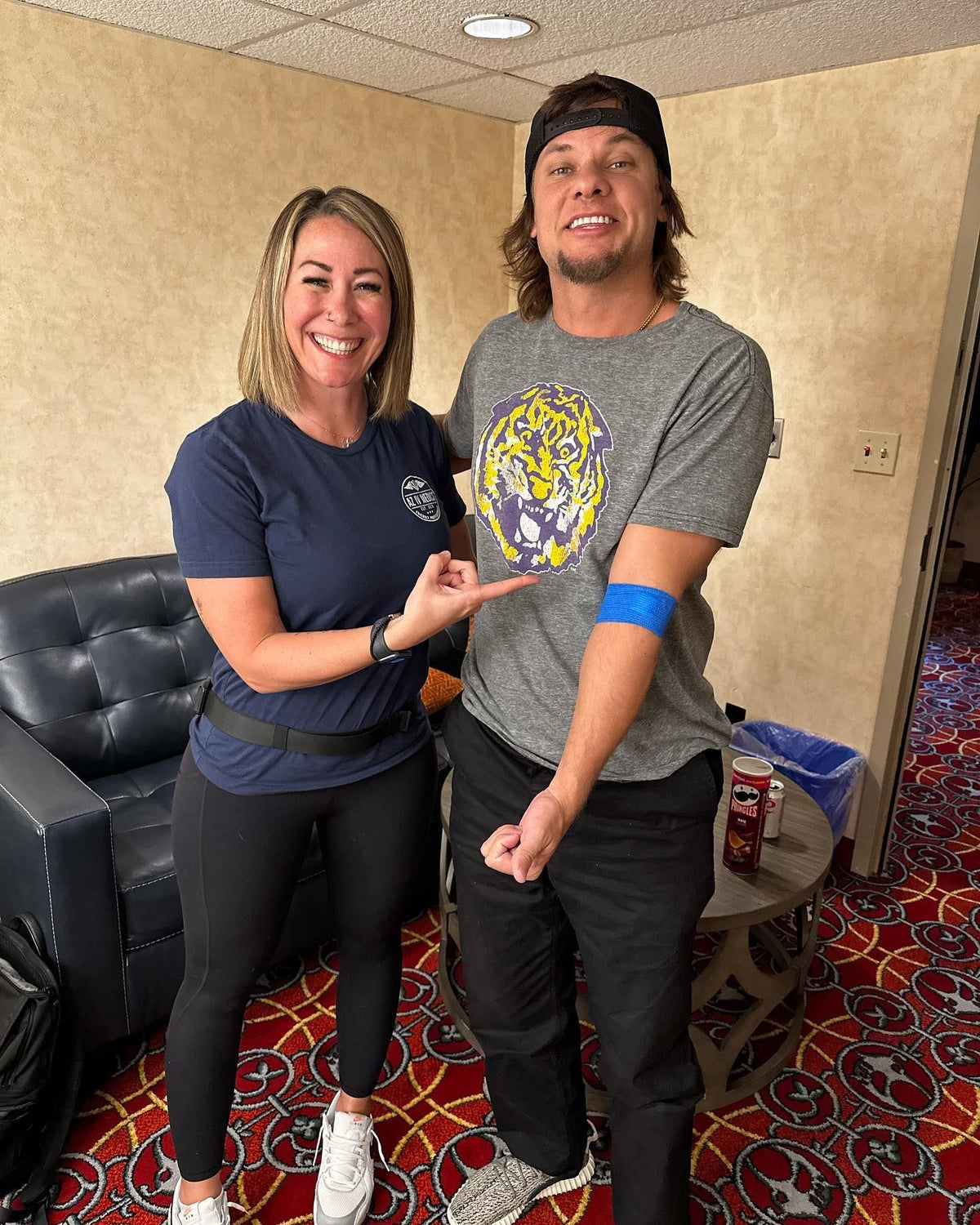 Woman points at man's bandaged arm. They smile, posing indoors near a chair. Man wears a shirt with a purple and gold logo.