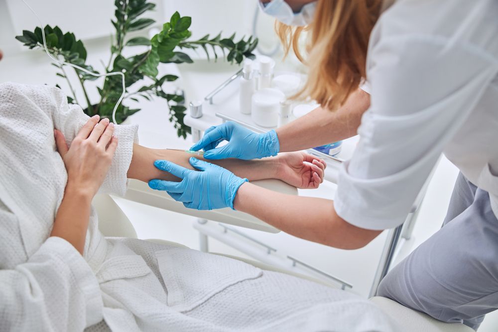 Person receiving intravenous treatment from a person wearing gloves and mask indoors.