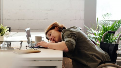 Person in a wheelchair asleep at a desk with a laptop, coffee mug, and plant.