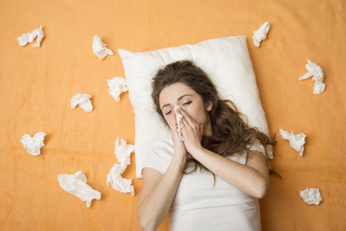 Woman in bed with a cold, blowing nose with tissues scattered on orange bedding.