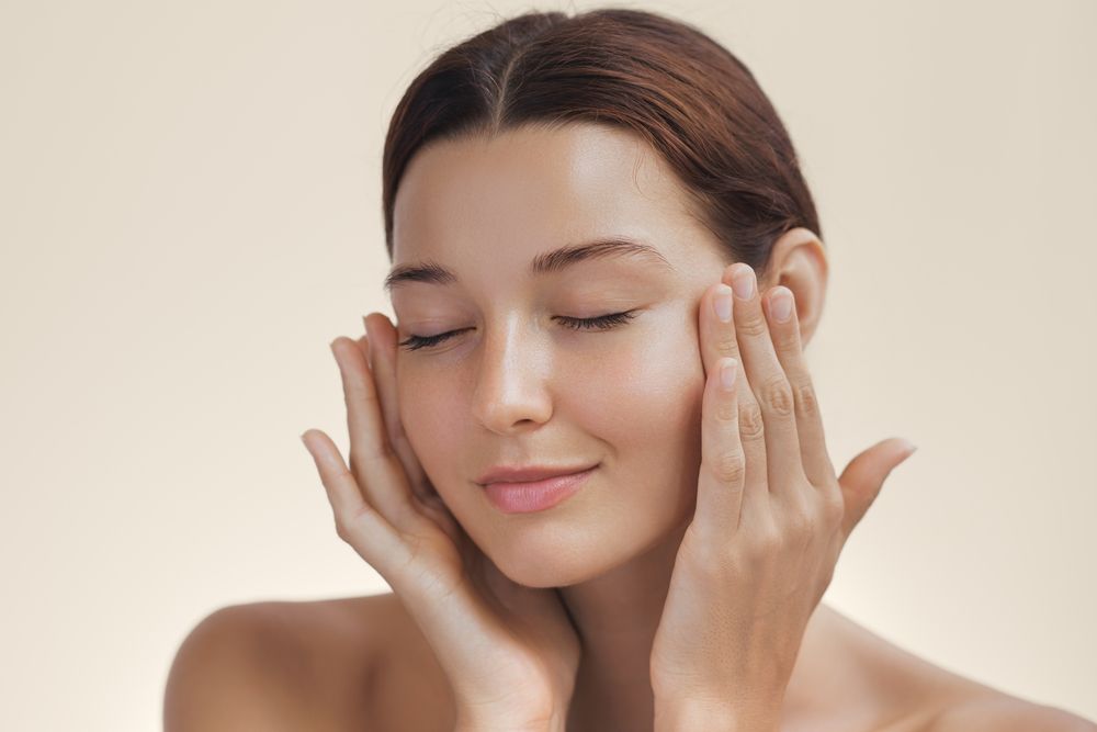 Woman with closed eyes, hands on face, soft focus, beige background.