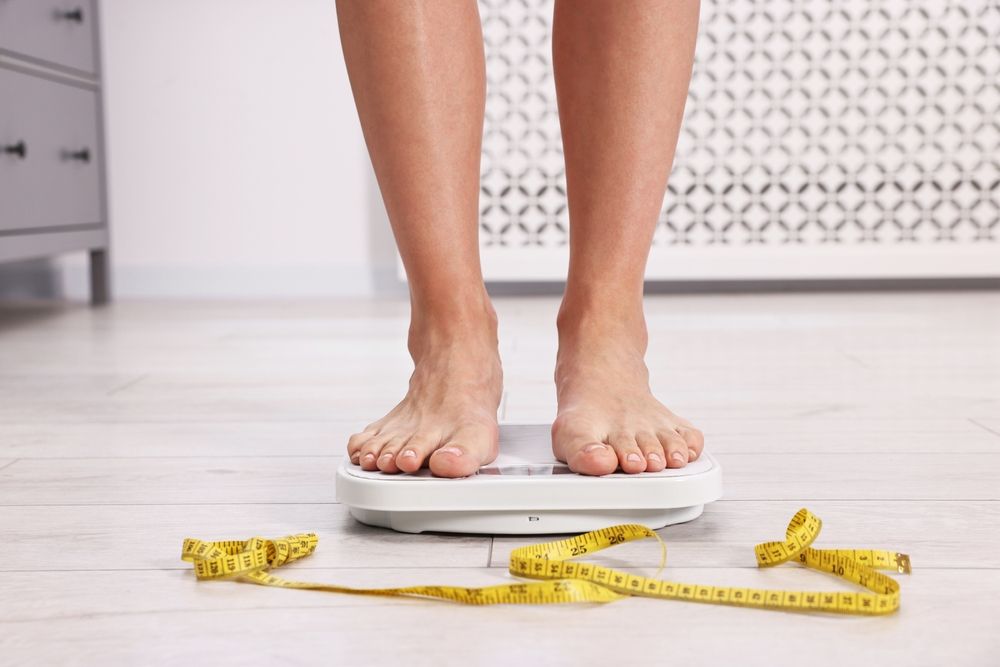 Bare feet standing on a weight scale; a yellow measuring tape lies on the floor.