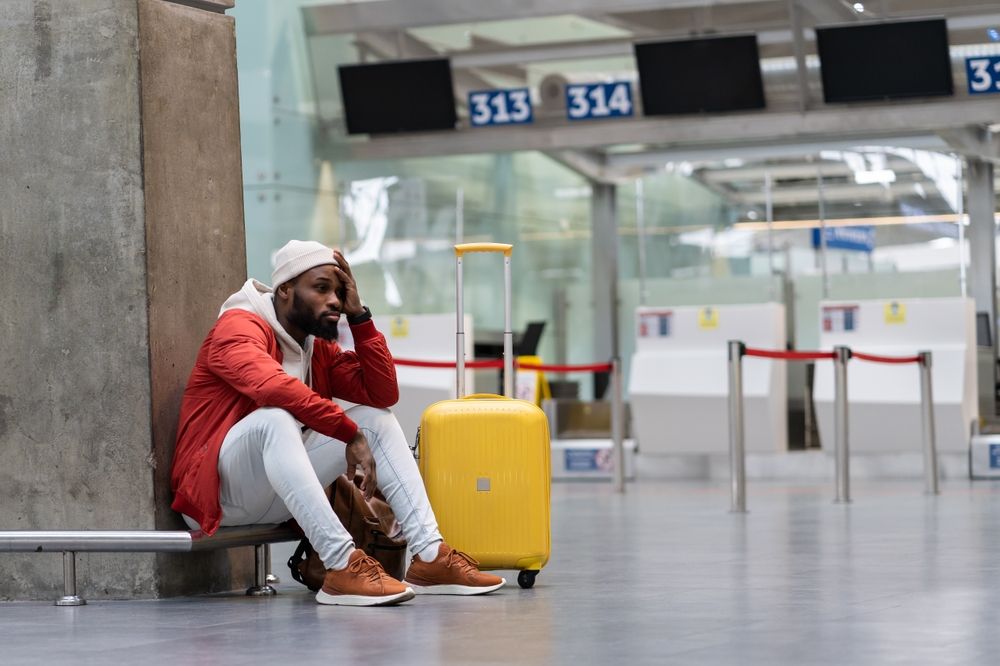 Man sits at airport, looking stressed, next to yellow suitcase.