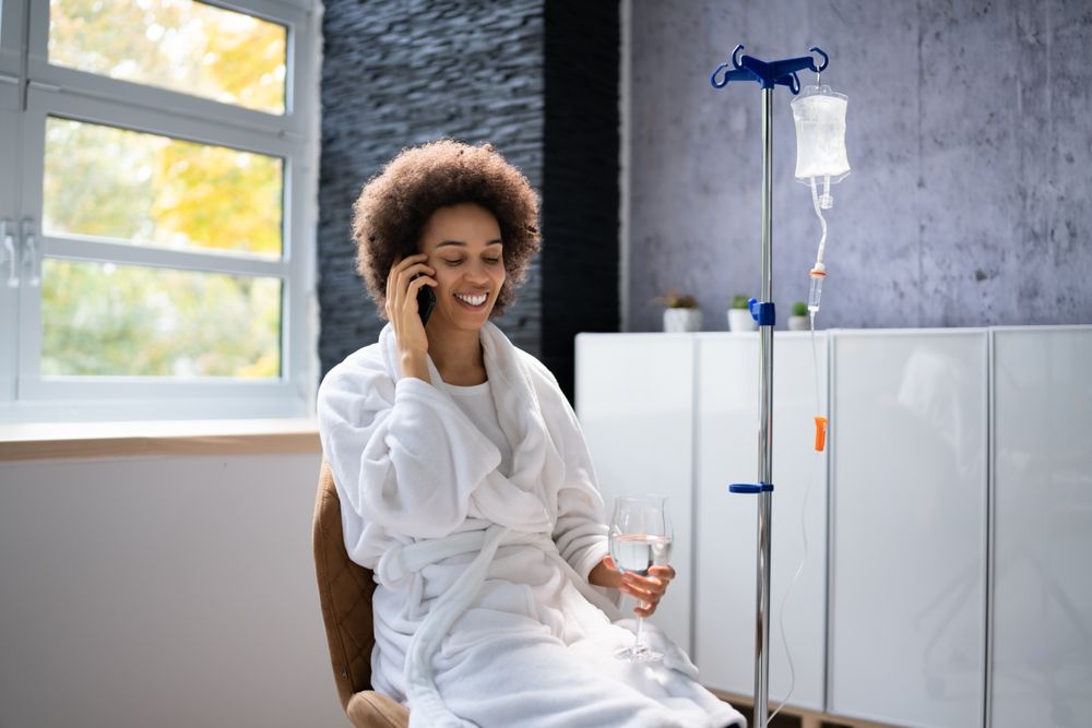 Woman in robe, receiving IV fluids, smiles while on a phone call. Bright room with window, white cabinet.