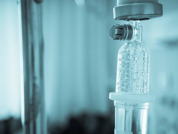 Close-up of an IV drip bag with liquid, attached to a medical pole in a pale blue-toned setting.