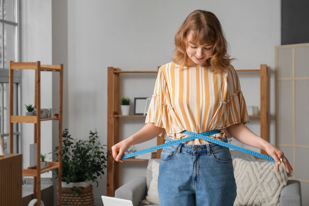 Woman measuring her waist with blue tape indoors, smiling.