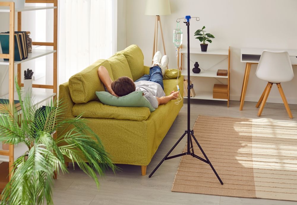 Person resting on sofa, receiving IV fluids at home, near a window and shelving.