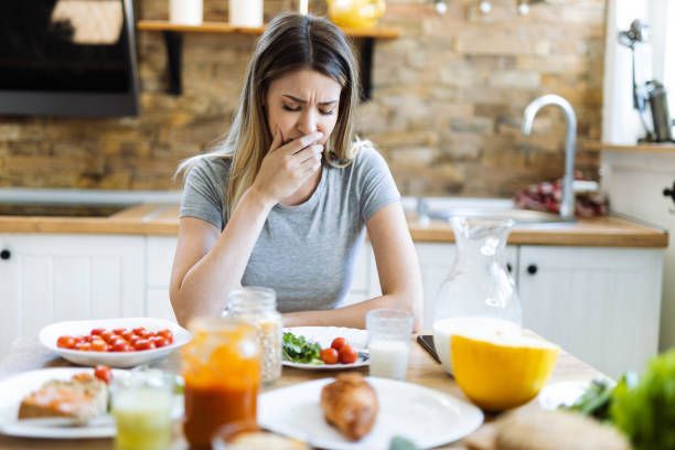 Woman in kitchen, appears nauseated. Hand covers mouth, food on table nearby.