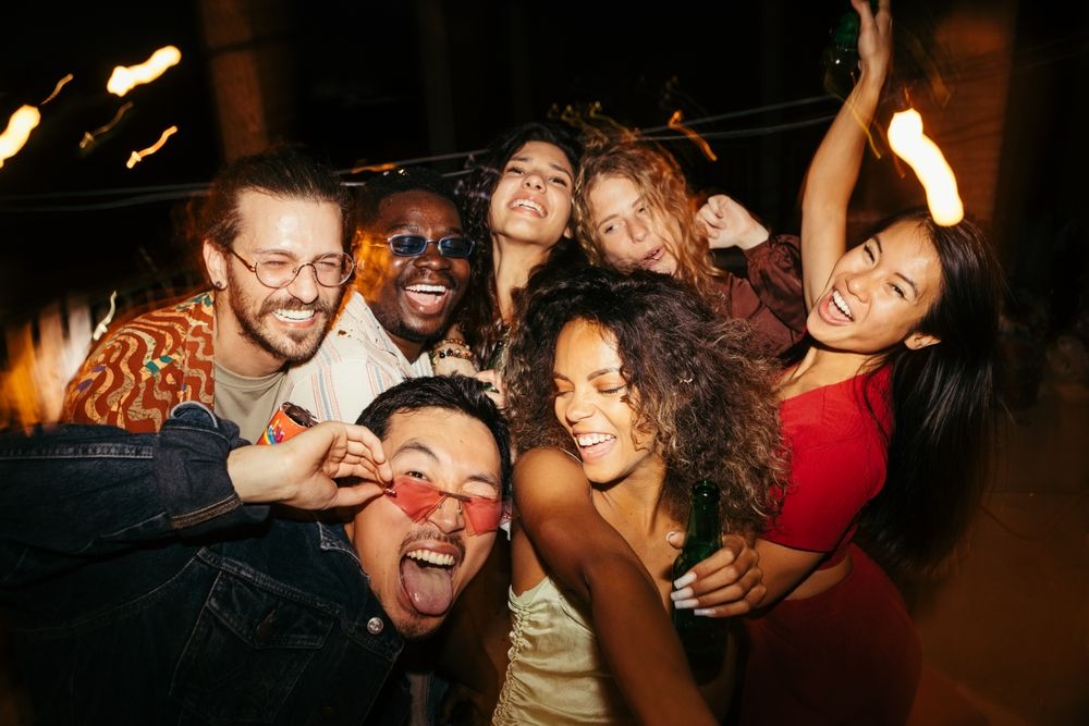 Group of friends laughing and smiling at a nighttime outdoor party; holding drinks, under string lights.