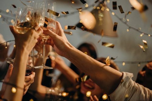 Hands clinking champagne glasses in a toast, gold confetti, lights, blurred faces, celebratory event.