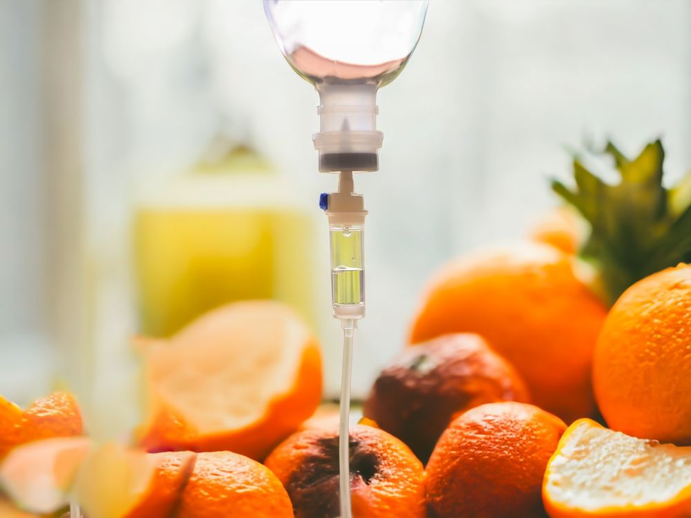 IV drip with clear fluid over a pile of oranges, some sliced. Bright, light background.