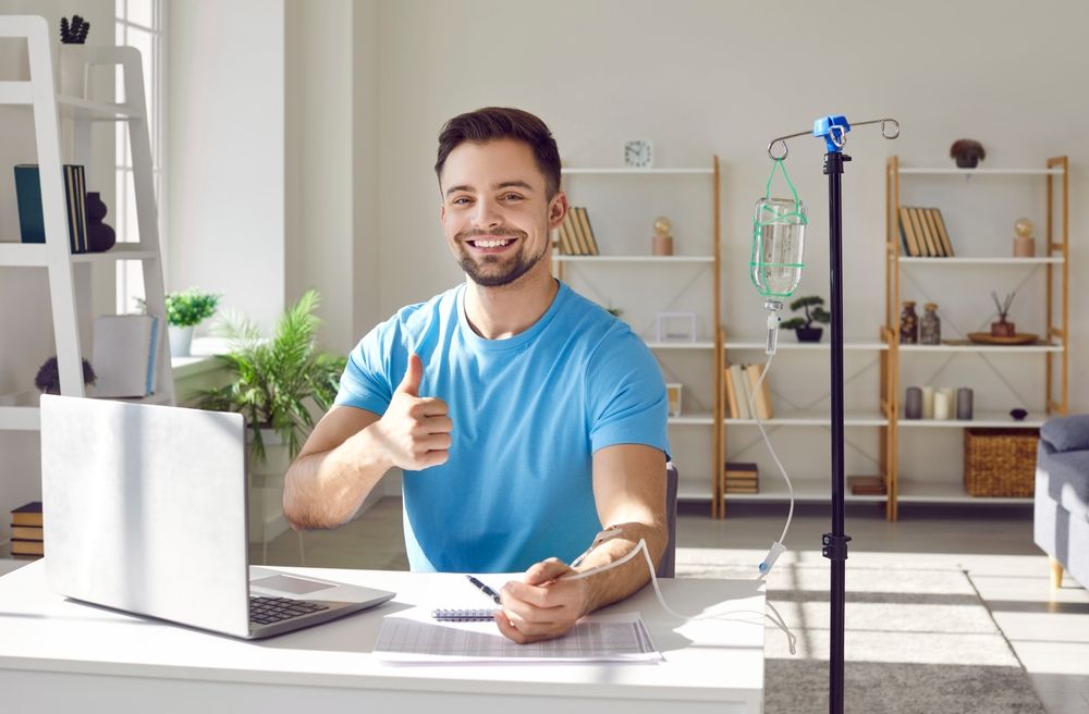 Man giving thumbs up while receiving IV fluids, sitting at a desk with a laptop.