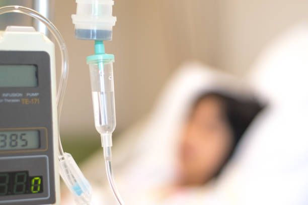 IV drip with clear fluid, and infusion pump, with a blurred person in a bed.