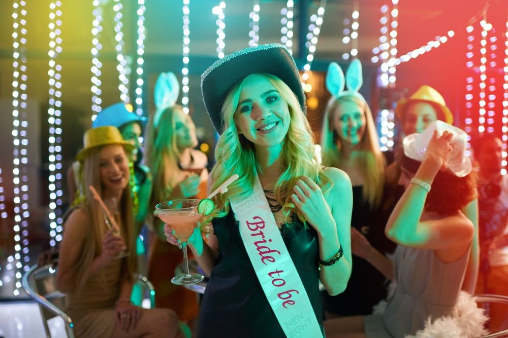 Bride-to-be at a bachelorette party, wearing a sash and cowboy hat, with friends in festive attire, holding a cocktail.