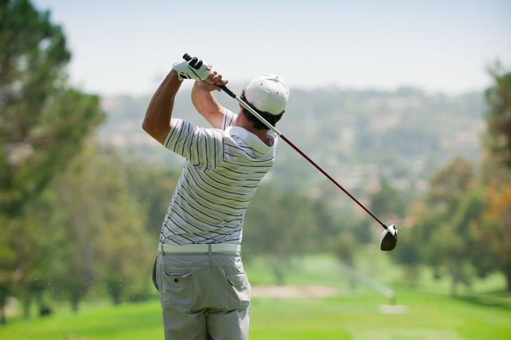 Golfer in a white hat and striped shirt swinging a club on a green golf course.